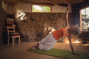 Frau mit weißerHose und orangenem Top in Yoga Haltung in einem keinen Yogastudio aus Naturmaterialien mit Holzboden und Naturlehm Wand